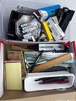 Top view of a plastic bin filled with black and yellow staplers, boxes of staples, some ink cartridges, multiple wooden and plastic rulers, pens, and adhesive note pads.