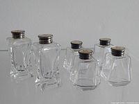 Group of six salt and pepper shakers with clear glass bodies and silver lids showing tarnish, arranged in two rows.