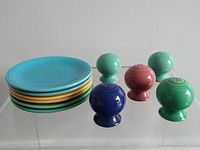 Stack of six saucers in turquoise, yellow, and orange colors alongside five round salt and pepper shakers in turquoise, blue, and pink colors