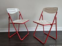 Two vintage folding chairs side by side, showing the red metal frame and white plastic seats and backs, highlighting condition and style