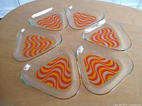 Six triangular glass hors d'oeuvres plates arranged on a round wooden table, showing the swirling op-art design in red, orange, yellow, and white.