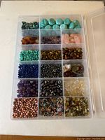Top view of a plastic compartment box containing 20 sections of various types of beads in different shapes, sizes, and colors including turquoise, amethyst, tiger's eye, and others.