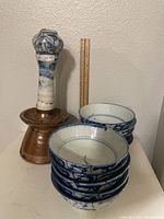 Photo shows six stacked blue and white Chinese porcelain bowls beside a tall ceramic candle holder with a brown base.