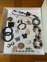 Top-down view of assorted costume jewelry displayed on white tray. Includes necklaces, bracelets, earrings, pendants, and rings with varied materials and styles. Black beaded pieces, gold-tone items, and butterfly themed pendants are visible.