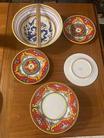 Photo showing one large hand-painted ceramic bowl and seven matching plates on a wood table including one plate face down revealing the 'Polic Italy' stamp