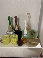 Multiple glass vases, tumblers, decorative items on a white surface against plain wall