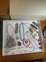 Top view of assorted vintage jewelry including necklaces, bracelets, brooches, clip-on earrings and a small silver mesh bag, arranged inside a white box.