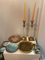 Photo showing entire lot with two lucite and brass candlestick holders with yellow candles and six enamel decorative plates in various colors and shapes.