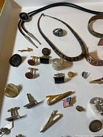 Men's accessories including bolo tie, rings, necklace, cufflinks, bracelets, tie clips, and pins spread out on white background in a box.