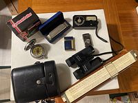 Overview of all items on wooden table: Car Dent Puller box, harmonica, compass, light meter, binoculars, slide rule
