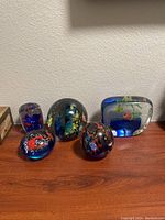 Front view of five different shaped glass paperweights with colorful designs embedded inside the glass, arranged on a shelf against a wall.