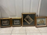 Four framed art pieces arranged on floor: two small architectural prints in matching gold frames, one larger framed print with a tilted black and white image, and one framed oil/enamel painting of a sailing ship.