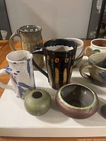 All ceramic items displayed on a surface: five mugs/cups and three small pottery bowls visible, with varied glaze styles.