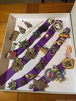 Wide view of all vintage brooches, pins, earrings, and necklace laid on purple ribbon inside a white display box.