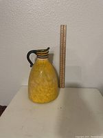 Front view of the yellow handblown glass pitcher with black handle next to a ruler showing about 9 inches height.