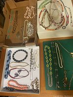 Top view photo showing multiple vintage necklaces laid out in cardboard boxes and on a green fabric surface. Various bead styles, chain types and colors visible.