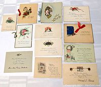 Group photo of eleven early Christmas cards spread on a flat surface showing front covers and greetings. Include various ribbons, printed seasonal messages and holiday imagery like flowers, lamps, cottages, and holly.
