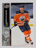 Front of Connor McDavid hockey card showing player in Edmonton Oilers orange uniform with black and white trim, holding hockey stick on ice rink.
