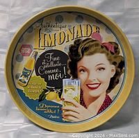 Photo of round metal serving tray with vintage french lemonade advertisement featuring a woman with brown hair holding a glass of lemonade against a yellow border.