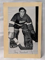 Front view of vintage Terry Sawchuck hockey goalie photo card in black and white showing the athlete robed in protective gear with hockey stick.