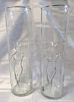 Pair of two tall clear cylindrical glasses with embossed nude female figure reliefs on each glass, standing side by side on white surface with soft lighting.