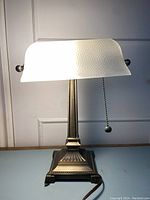 Front view of the brass banker's lamp with frosted embossed glass shade illuminated, showing heavy solid metal base and decorative detailing