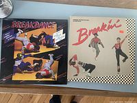 Two 1980s breakdance vinyl LP records cover art side by side on floor. One album titled 'Breakdance' features four photos of breakdancers in various poses. Other album titled 'Breakin'' shows three breakdancers with checkered border.