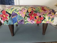 Front view of small footstool covered in colorful floral and geometric patterned fabric with four dark tapered wooden legs.
