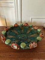 Front angled photo showing the blue-green ceramic dish with frogs and lily pads around the rim, highlighting the colors and sculpted details on each figure.