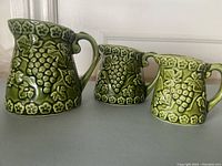 Three green ceramic measuring cups of varying sizes with grapevine motif, arranged side by side