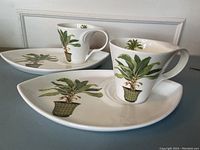 Two sets of leaf-shaped porcelain snack plates and teacups with detailed handpainted palm plant designs. Cups have a palm plant inside and outside, plates have palm plants on surfaces.