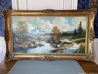 Full front view of the framed landscape oil painting showing the composition of mountains, river, trees, and sky.