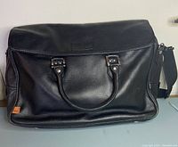Front view of black leather messenger bag showing top handles and front flap closure with metal hardware.
