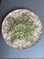Full view of circular decorative china plate with painted Mountain Avens flowers and foliage on crackled beige background with gold rim.