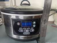 Front view of Hamilton Beach slow cooker showing digital control panel and stainless steel housing with black accents, a measuring ruler leaning beside it to show size.