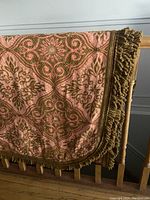 Bedspread hanging over wooden railing showing pink and brown/gold floral and swirl pattern with thick fringe on three sides.