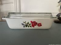 Side view of white rectangular milk glass Pyrex casserole featuring red rose floral design and green leaves with clear glass lid in place.