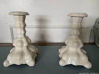 Two cream-white ceramic candlestick holders with ornate sculpted bases and wide tops, glossy finish.