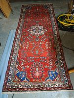 Full view of the runner carpet laid on concrete floor showing red field, central medallion, floral motifs, ivory guard borders, blue corner accents and fringed ends