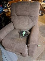 Brown fabric La-Z-Boy power recliner showing the chair's full front, armrests, and cushioned backrest. A plastic bag containing a remote control labeled 'Recliner' rests on the seat.