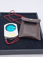 Photo showing the clinometer device and brown leather carrying pouch on black background.
