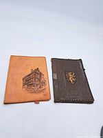 Two leather folio covers side by side on white background; left tan with building drawing, right dark brown embossed with gold crest.