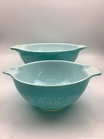 Photo showing front view of two turquoise and white vintage Pyrex bowls with folk art pattern and dual handles