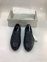 Front view of the black quilted slip-on GEOX shoes with tag attached and box in background.
