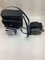 Photo showing both cameras and the black leather carrying case with shoulder strap, set on white background.