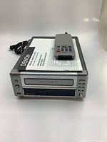 Front view of the Denon tape recorder showing the cassette slot, front control buttons, power cable, included remote, and instruction manual on top.