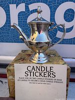 Front view of the silver tone metal teapot container with hinged lid and spout, showing the reflective surface and ornate handle.