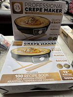Box of the Professional Crepe Maker with details about non-stick aluminum surface, included utensils, recipe book, and power specs.