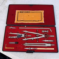 Wide view of the open drafting set case showing all drafting tools, compass parts, adaptors, and the yellow cloth with Dietzgen Stellar branding.