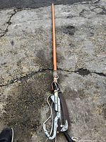 Full length view of the orange Fiskars pole saw lying on rough concrete, showing the entire pole and saw blade.